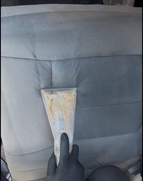 Hand in black glove scraping dirty substance off white quilted car seat with plastic scraper tool