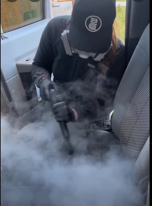 Person in black hoodie sitting in car seat surrounded by vapor or fog from vaping device